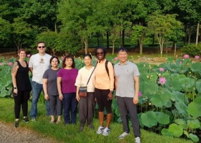 Six people standing in front of a pond with floewring plants and trees in the background