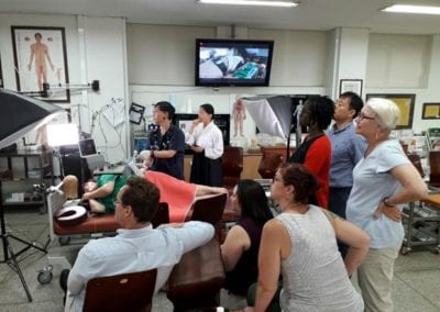 Eight people around an table with someone laying down and group observing a screen that appears to be out of frame