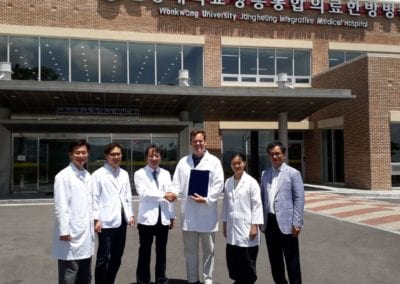 Six doctors standing in front of medical hospital. Middle two shaking hands and one holding a certificate.