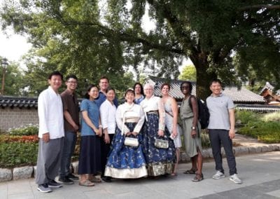 Group of people standing under a tree. Two women in middle wearing traditional dresses.