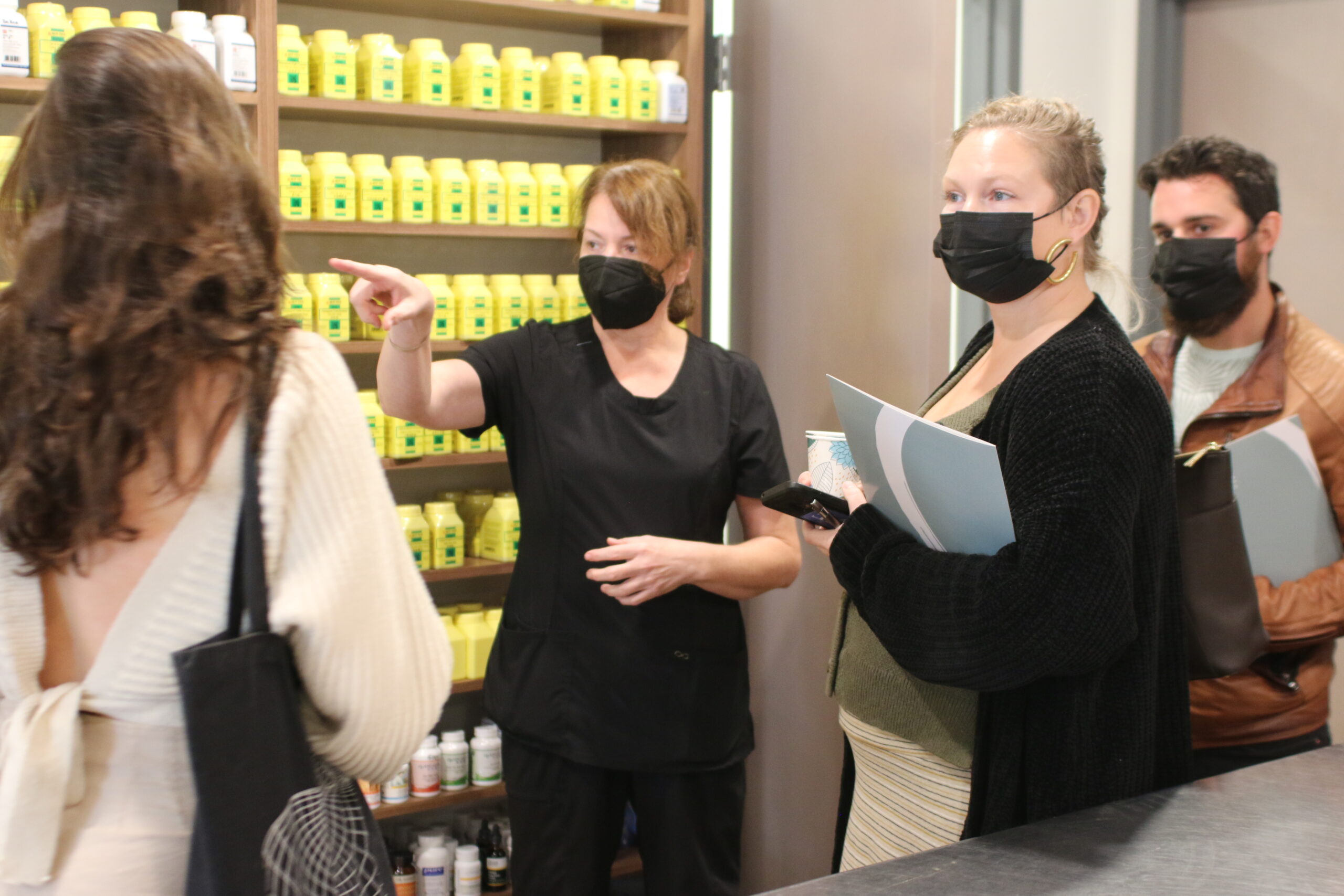 a woman wearing a black mask points to a shelf full of yellow bottles
